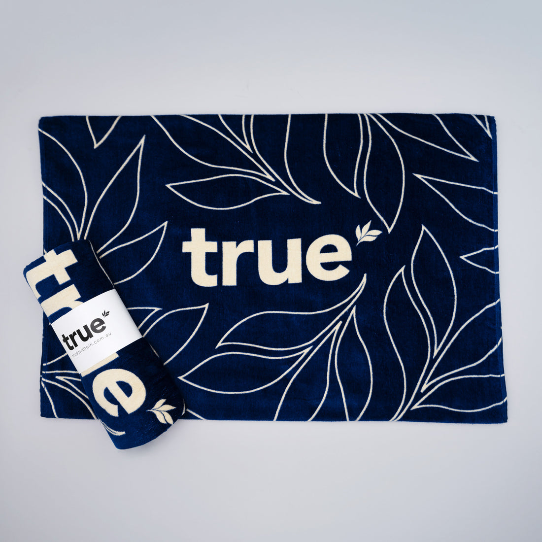 Navy blue &quot;true&quot; beach towel with white botanical leaf print and logo, rolled and laid flat — cotton beach towel product photo for travel, beach, and pool.