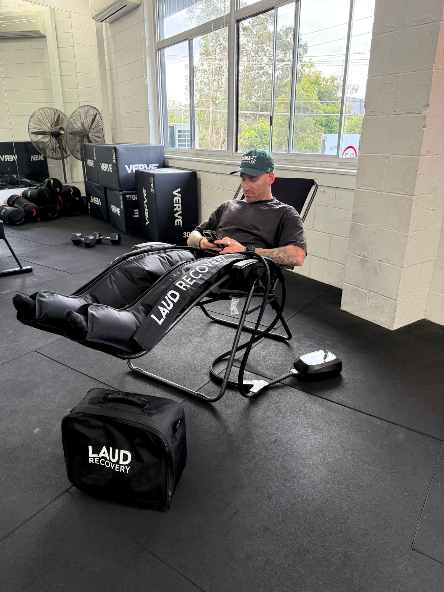 Man reclined in a gym recovery area using LAUD Recovery black compression leg boots with control unit, holding phone; gym equipment and stacked VERVE plyo boxes visible in background.