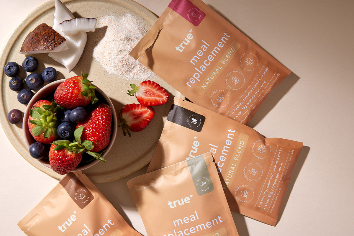 True meal replacement natural blend sachets on a beige countertop with a bowl of strawberries and blueberries, coconut pieces and powdered shake — plant-based meal replacement protein shake packets for healthy breakfast and weight management.