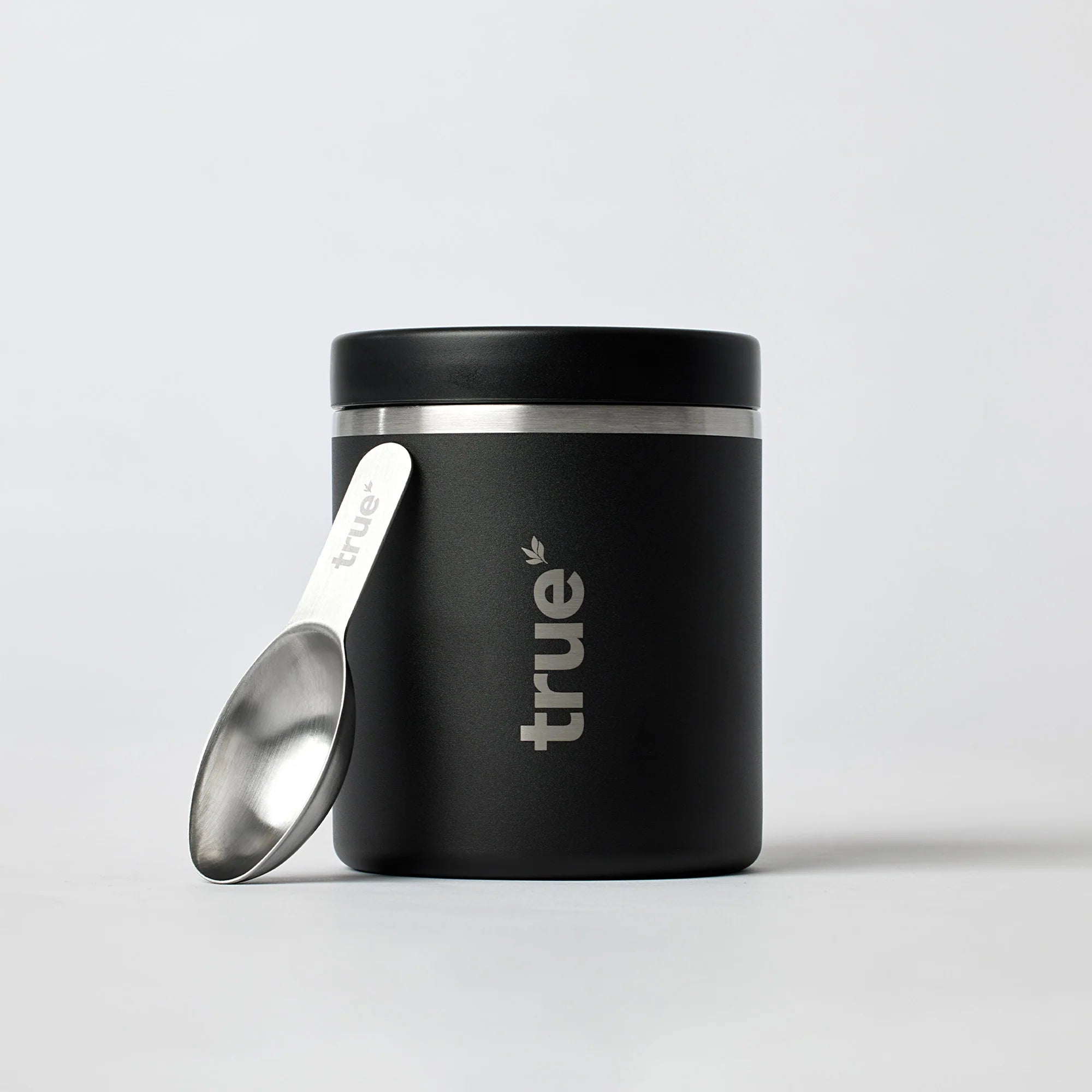 Black insulated stainless steel food jar with silver lid trim and matching stainless scoop leaning against it, branded "true" on front — minimalist product shot on light grey background.