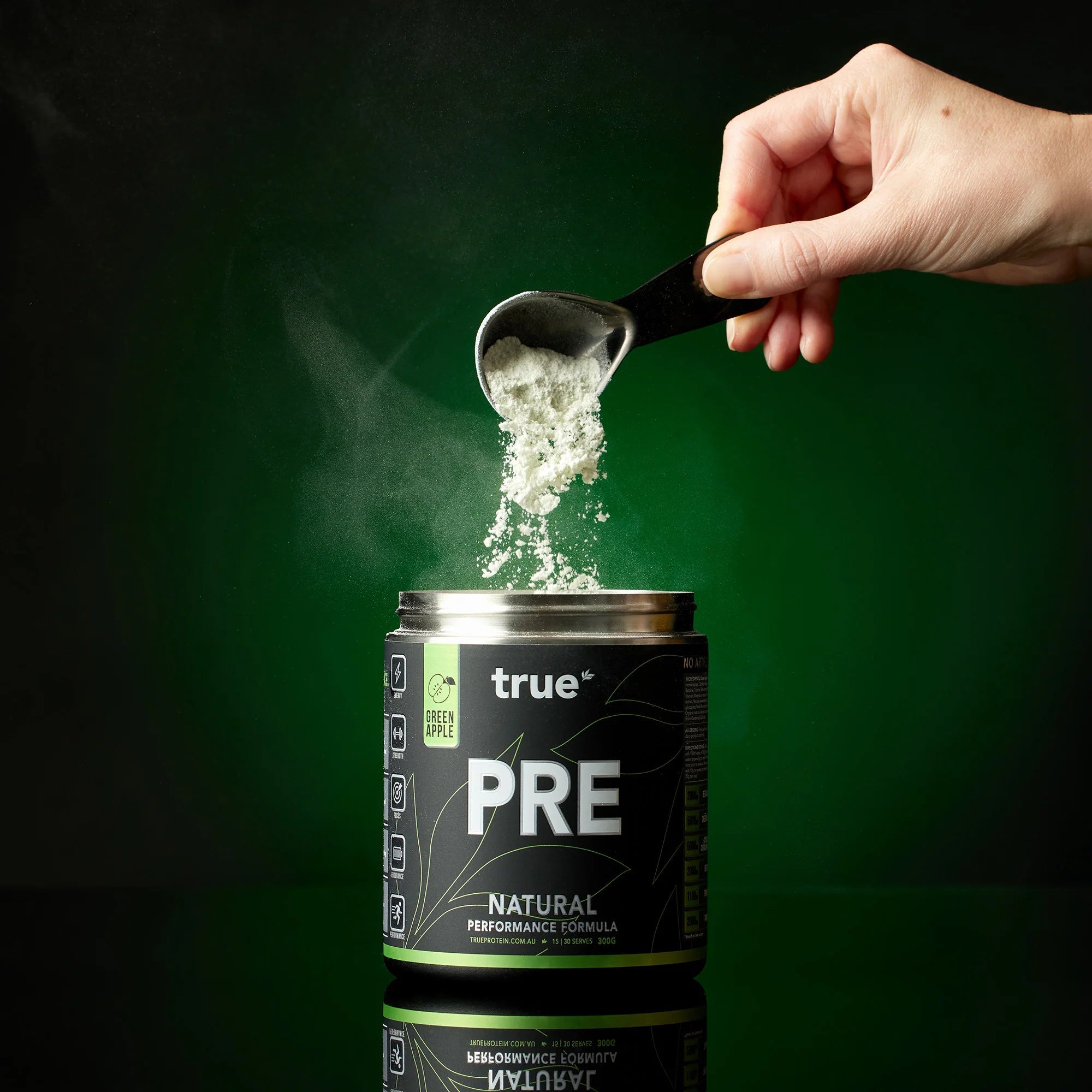 Close-up studio shot of True PRE natural pre-workout powder (green apple flavor) being scooped above an open black-green tub on a dark green background — supplement product image for fitness and performance.
