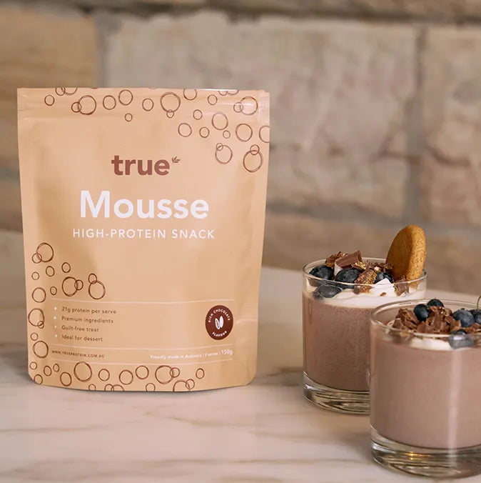 True Mousse high-protein snack powder pouch beside two chocolate mousse desserts in glasses topped with whipped cream, blueberries, chocolate shavings and a biscuit — True brand protein mousse made in Australia.