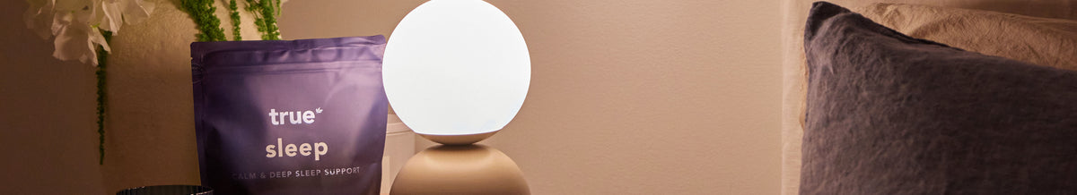 A True Sleep sleep aid supplement pouch in purple packaging next to a glowing spherical lamp on a bedside table, with a bed and flowers in a softly lit bedroom setting.