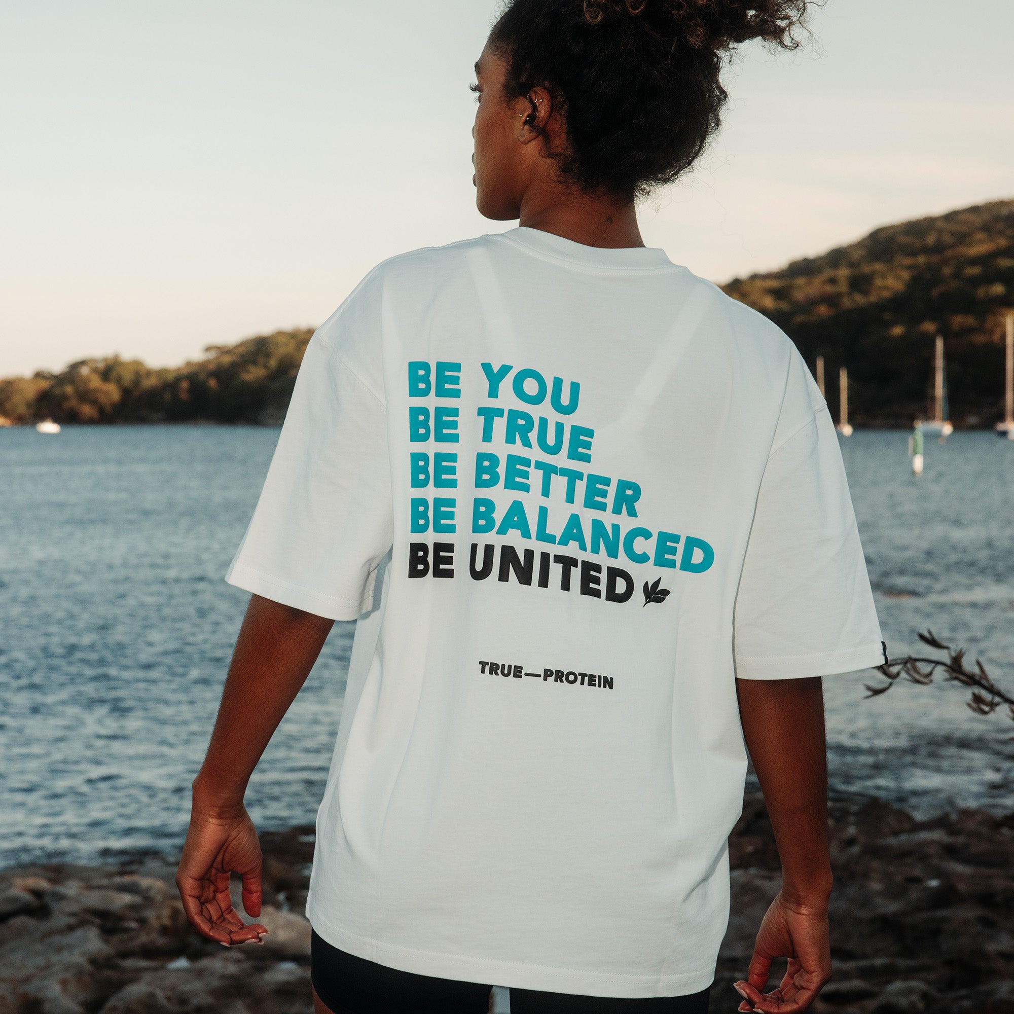 Unisex oversized white cotton tee with bold turquoise and black back graphic reading "BE YOU BE TRUE BE BETTER BE BALANCED BE UNITED" and small "TRUE—PROTEIN" logo, worn by a model in a coastal waterfront lifestyle scene with sailboats and rocky shoreline.