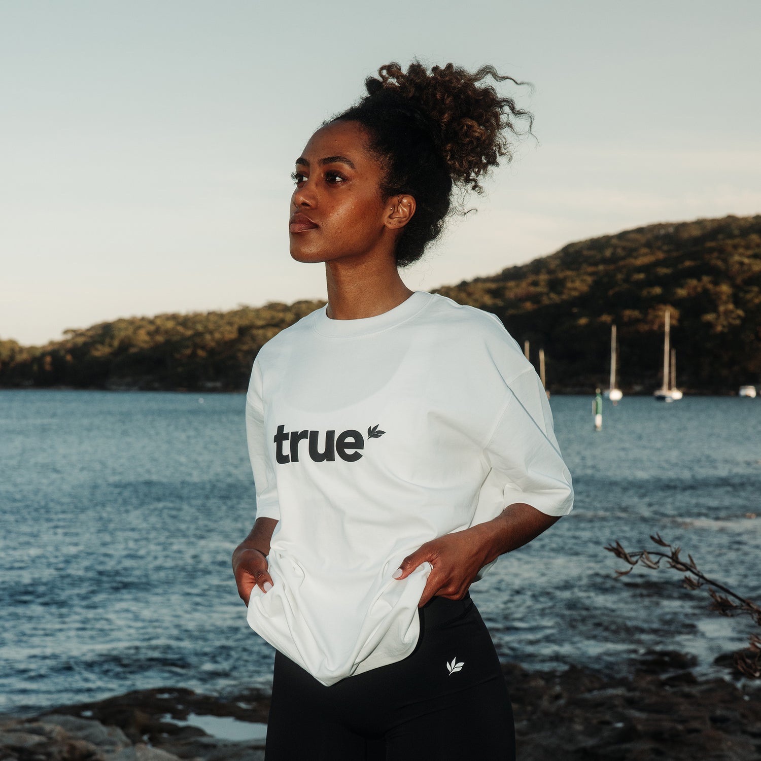 White unisex oversized tee with black "true" logo worn by a woman model standing at a coastal harbor with sailboats in the background — lifestyle product shot for casual eco-friendly clothing.