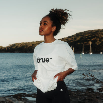 White unisex oversized tee with black "true" logo worn by a woman model standing at a coastal harbor with sailboats in the background — lifestyle product shot for casual eco-friendly clothing.