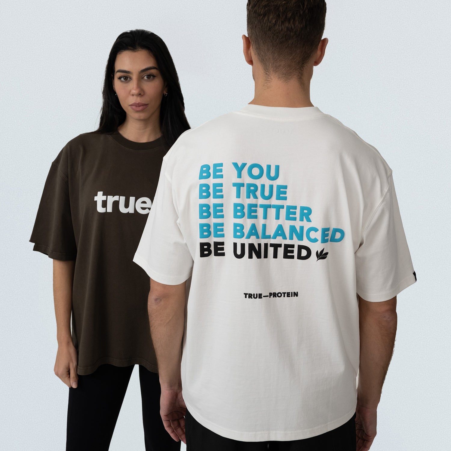 Unisex oversized cotton t‑shirts — brown front‑view tee with white "true" logo and white back‑view tee printed in blue/black text "BE YOU BE TRUE BE BETTER BE BALANCED BE UNITED" with "TRUE_PROTEIN" brand logo, studio product shot for online apparel store.