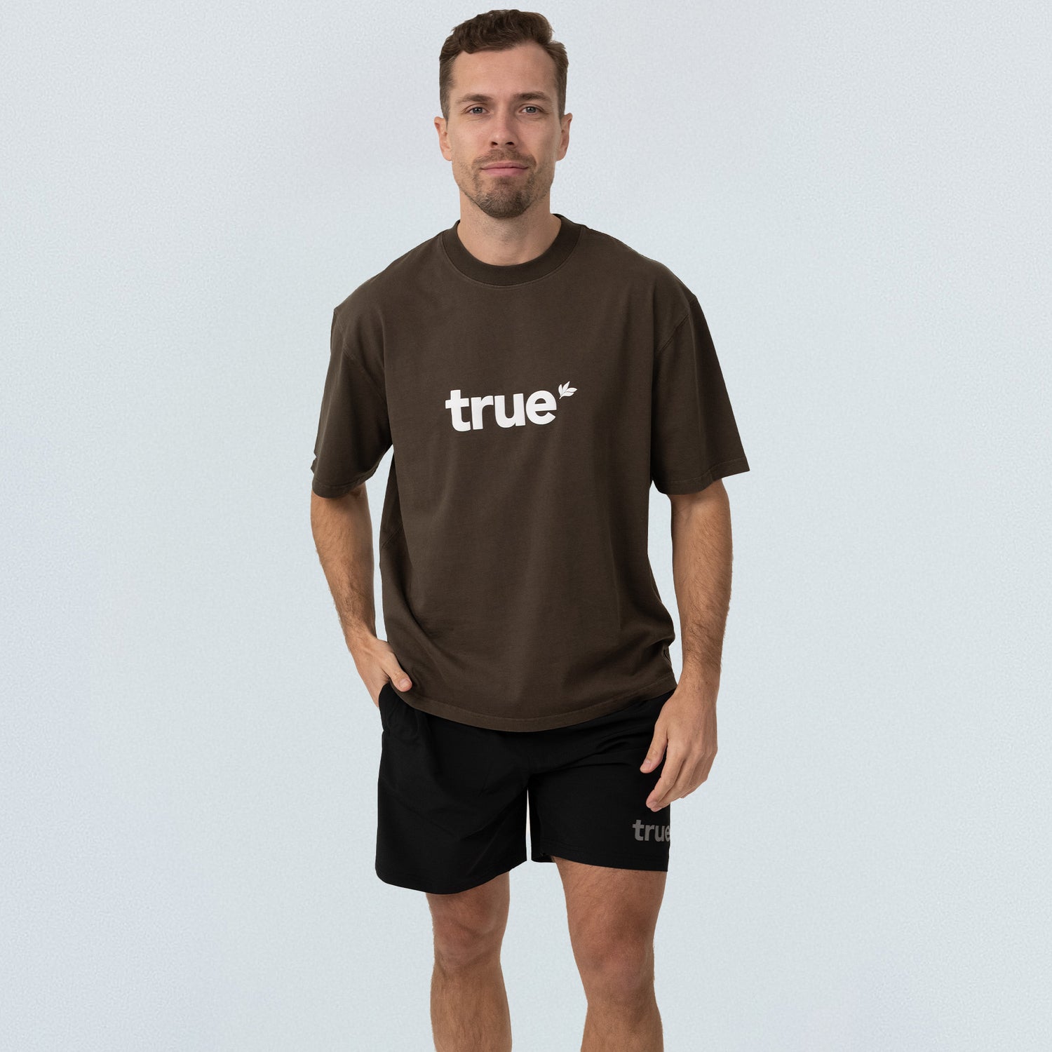 Male model in brown oversized unisex cotton T‑shirt with white "true" logo and black "true" shorts on light studio background — casual streetwear product shot, front view.