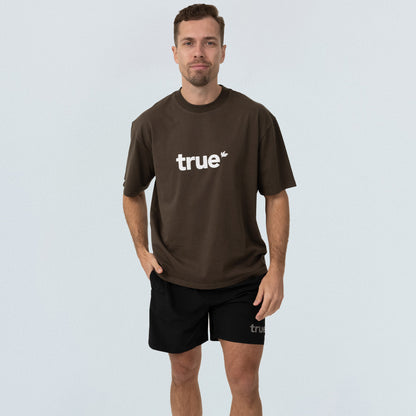 Male model in brown oversized unisex cotton T‑shirt with white "true" logo and black "true" shorts on light studio background — casual streetwear product shot, front view.