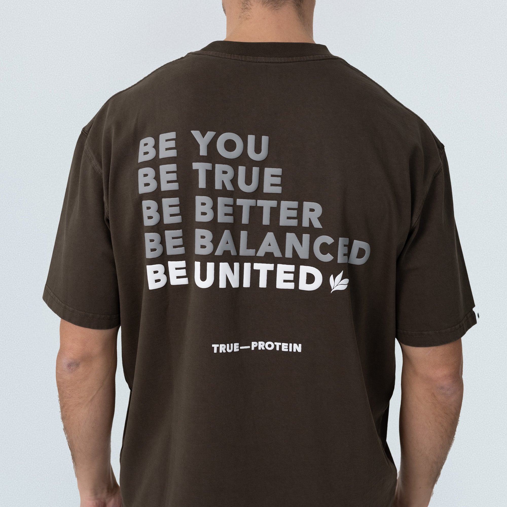 Unisex oversized dark brown cotton t-shirt, back view on model, white block print reading “BE YOU BE TRUE BE BETTER BE BALANCED BE UNITED” with small “TRUE—PROTEIN” logo at lower back — casual streetwear tee product photo.