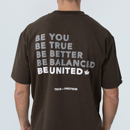 Unisex oversized dark brown cotton t-shirt, back view on model, white block print reading “BE YOU BE TRUE BE BETTER BE BALANCED BE UNITED” with small “TRUE—PROTEIN” logo at lower back — casual streetwear tee product photo.