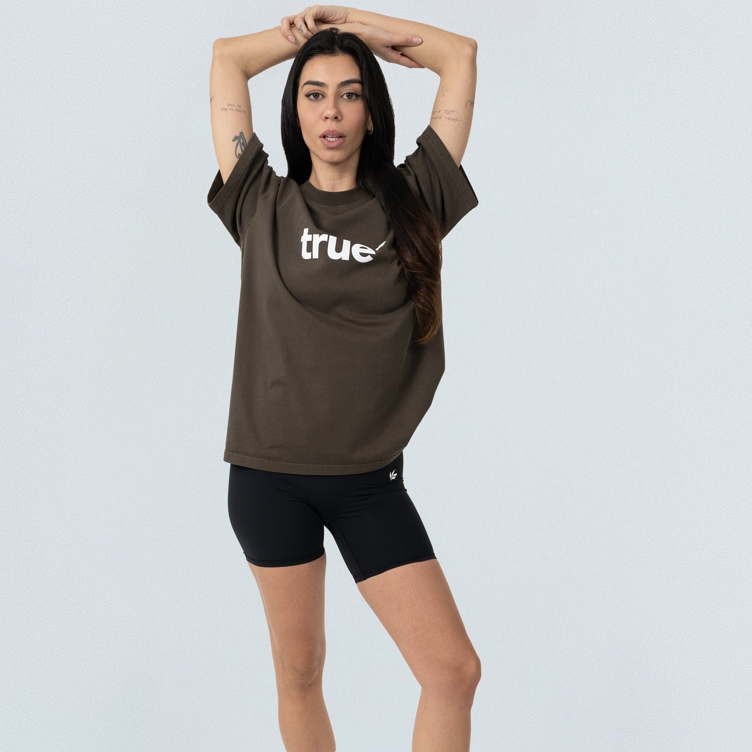 Olive-brown unisex oversized "true" logo tee on model, paired with black bike shorts, front view on light studio background — relaxed-fit casual streetwear.