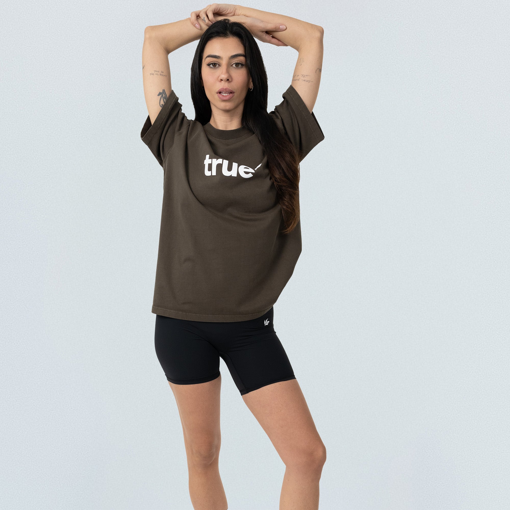 Olive-brown unisex oversized "true" logo tee on model, paired with black bike shorts, front view on light studio background — relaxed-fit casual streetwear.