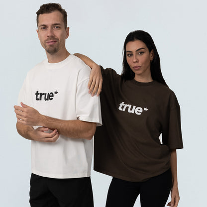 Two models wearing unisex oversized cotton tees with "true" logo — man in white tee and woman in dark brown tee, studio shot, relaxed fit casual streetwear product image.