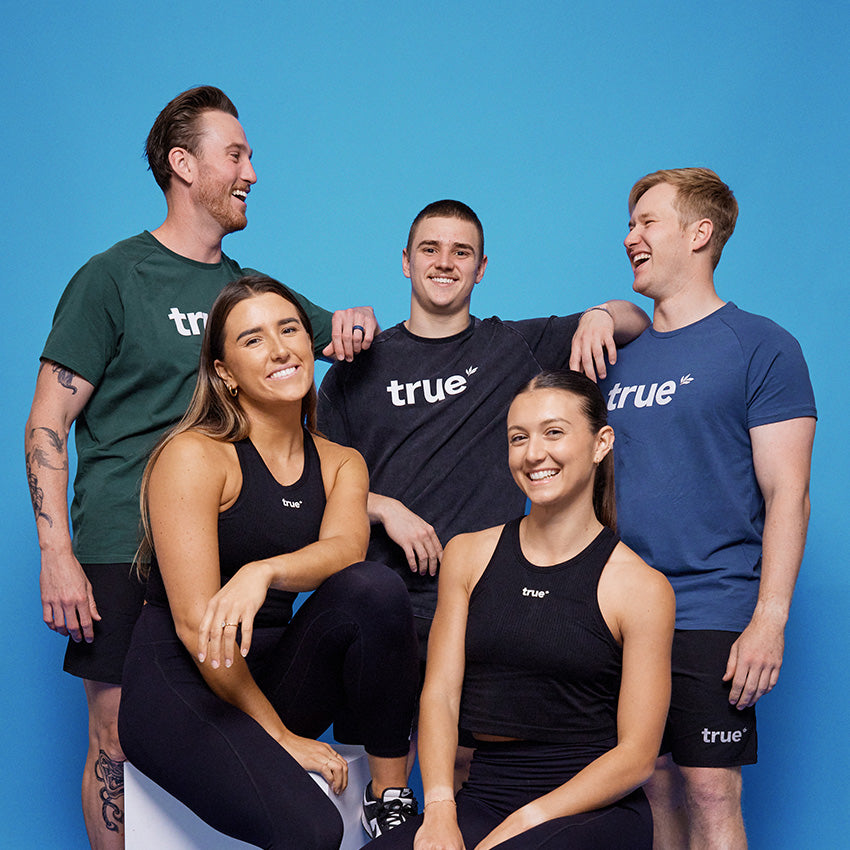 Five smiling young adults wearing True branded activewear (tank tops, tees, shorts) posing in a studio portrait with a solid blue background — lifestyle photo for True activewear and group fitness apparel.
