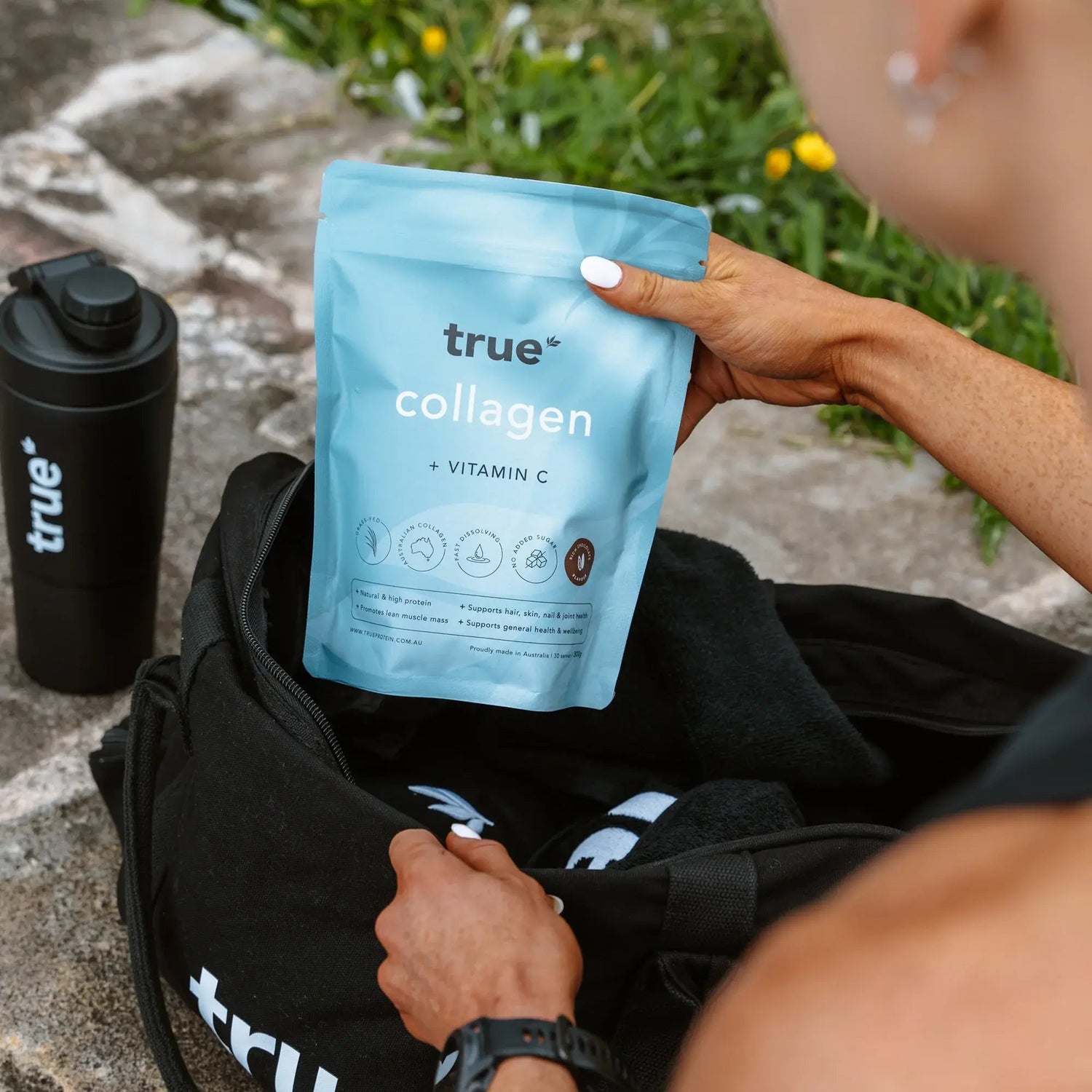 True Collagen + Vitamin C powder pouch (True brand) held above a black gym bag with matching True shaker bottle outdoors — fitness supplement product photo, Australia-focused SEO.