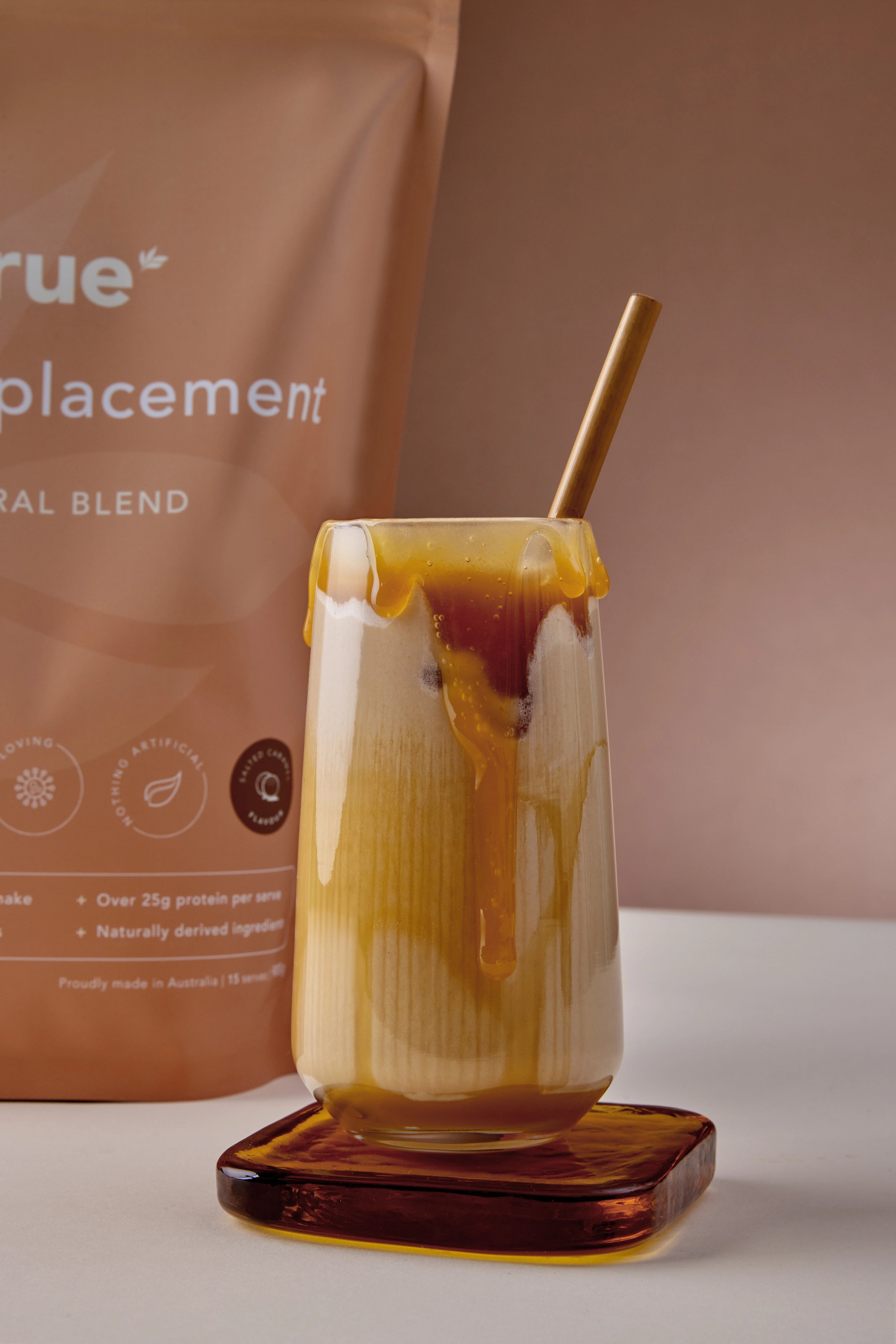 Glass of salted caramel meal replacement shake with caramel drizzle and bamboo straw beside True Protein pouch, naturally derived ingredients, proudly made in Australia—product shot for fitness nutrition.