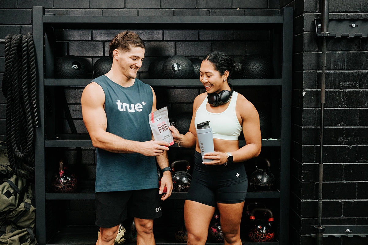 Two people holding bottles in a gym setting with black equipment in the background - True Protein Non-stim Pre