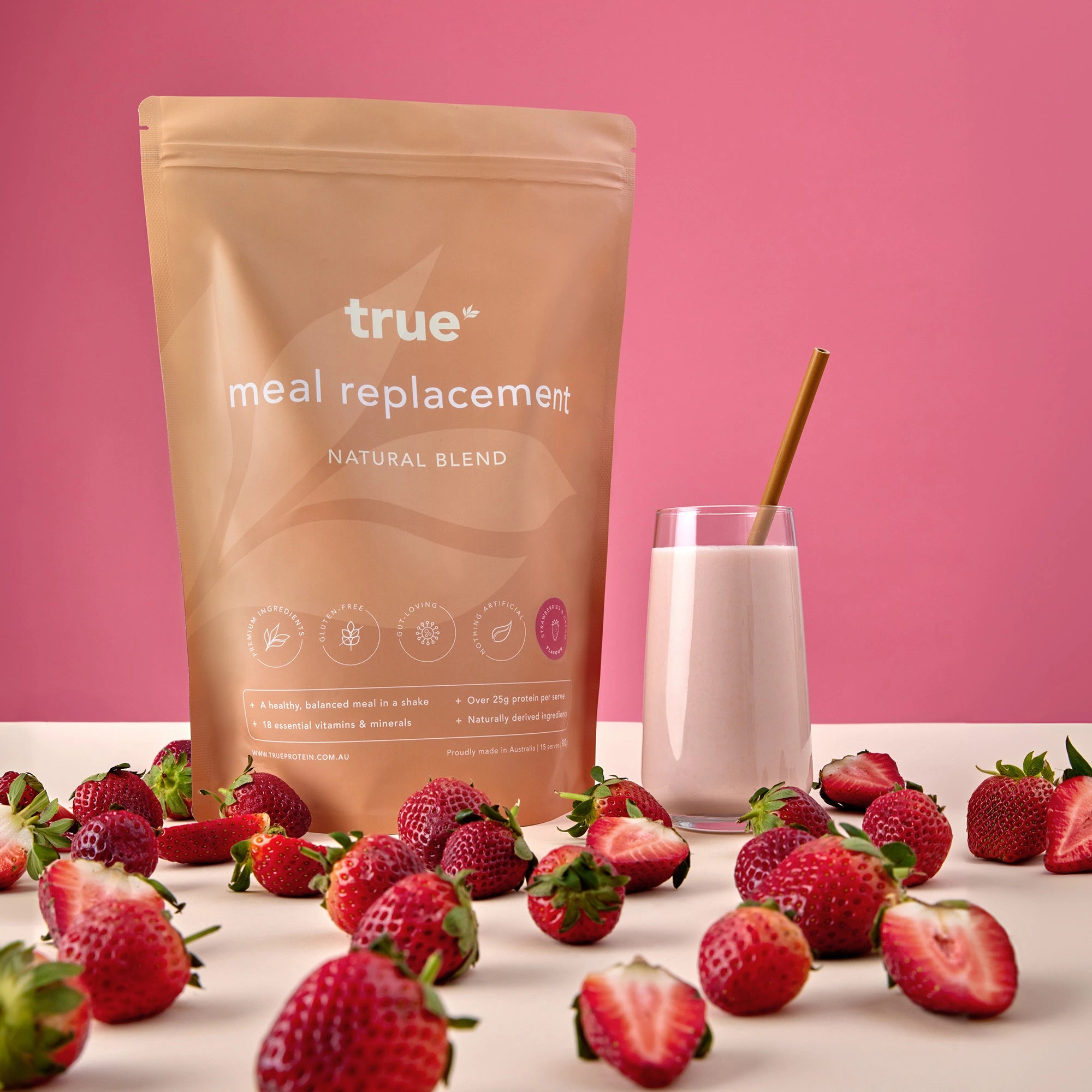 True meal replacement natural blend pouch (True Protein) beside a strawberry protein shake in a glass with straw, fresh strawberries scattered on a pink background — Australian-made meal replacement powder, strawberry flavor.