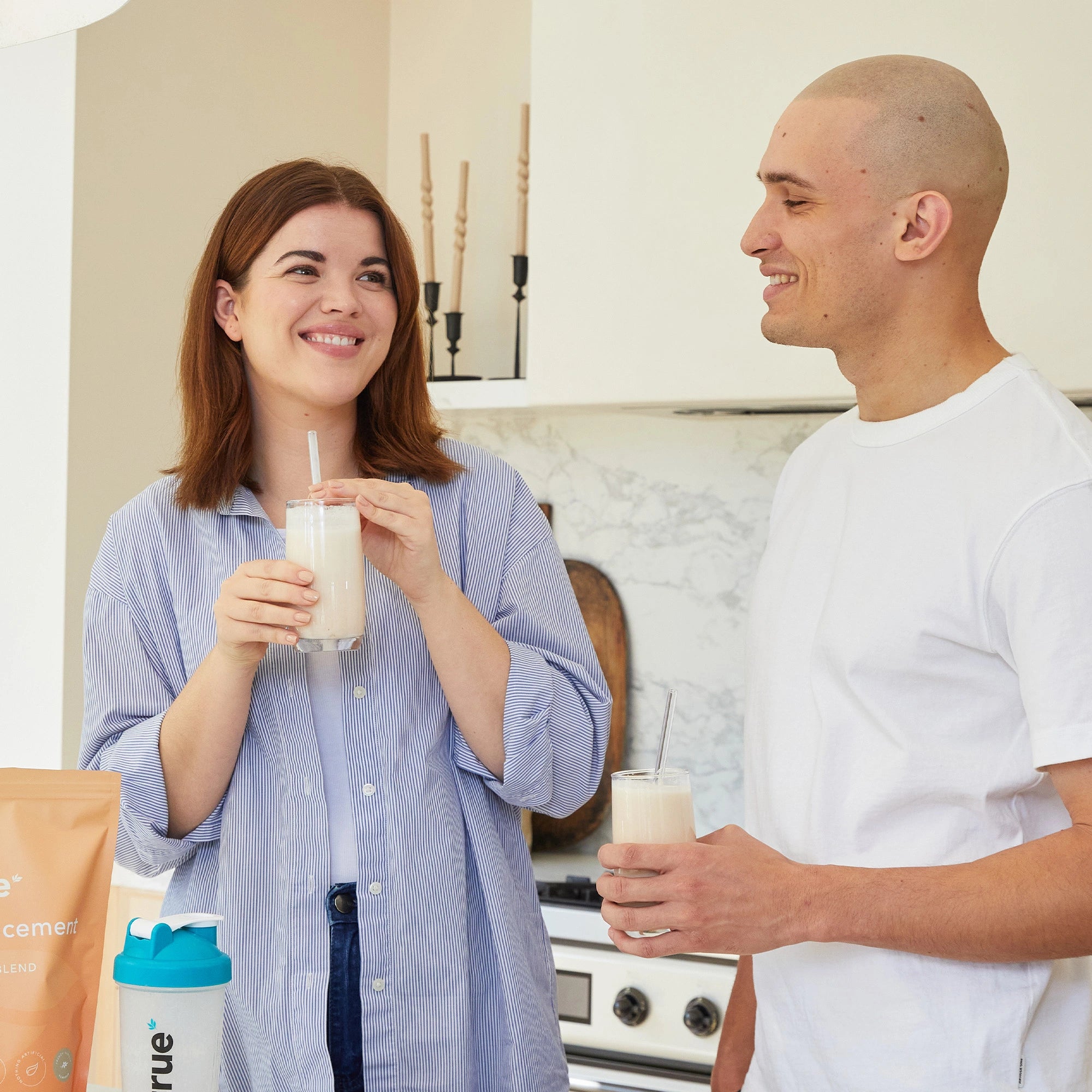 Two adults in a modern home kitchen smiling and holding creamy meal-replacement protein shakes with a True Protein bag and branded shaker visible on the counter — healthy protein shake breakfast lifestyle.