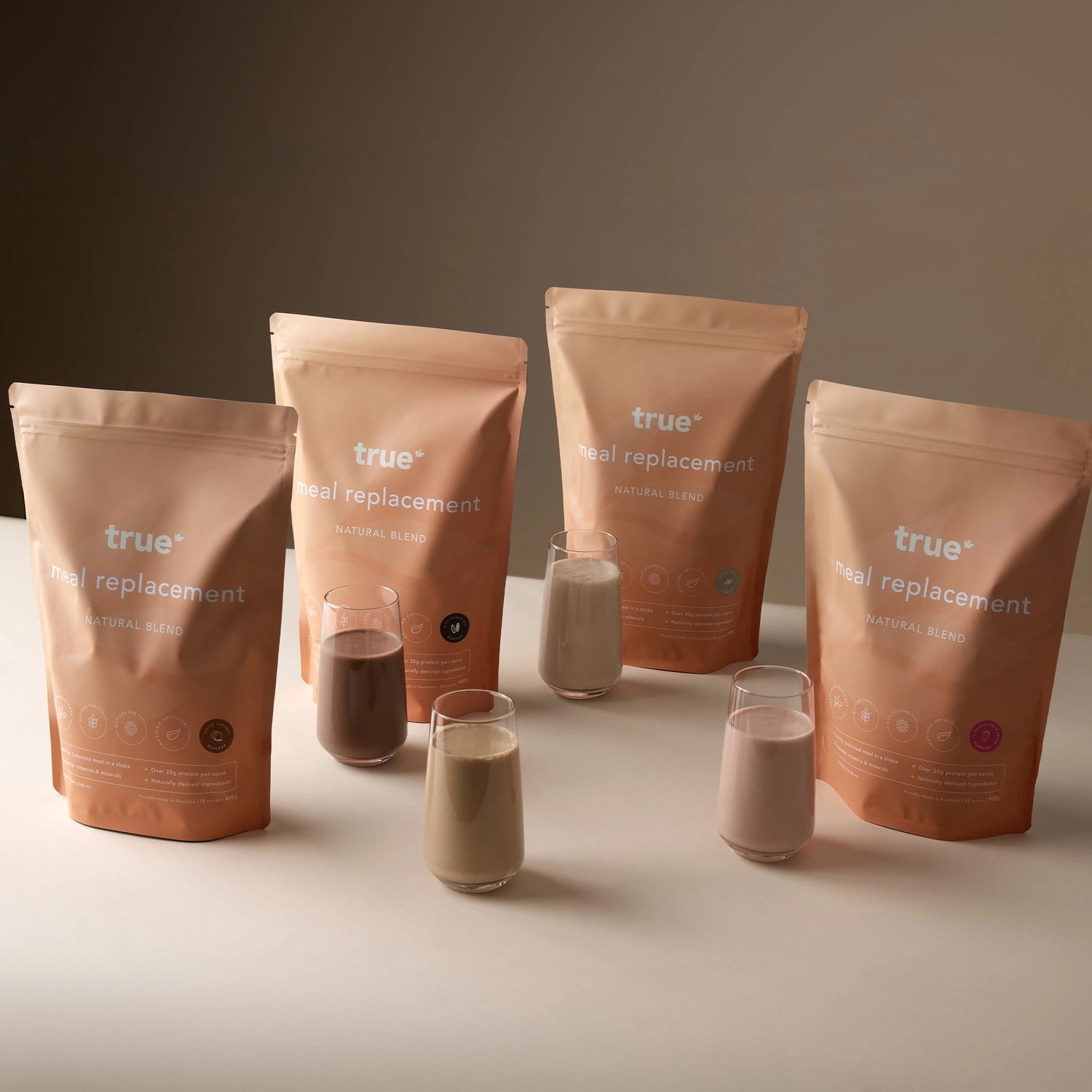 True Meal Replacement Natural Blend resealable pouches (four) with four prepared protein shakes in glasses on a neutral beige background — meal replacement powder, protein shake pouch, healthy shake product photo