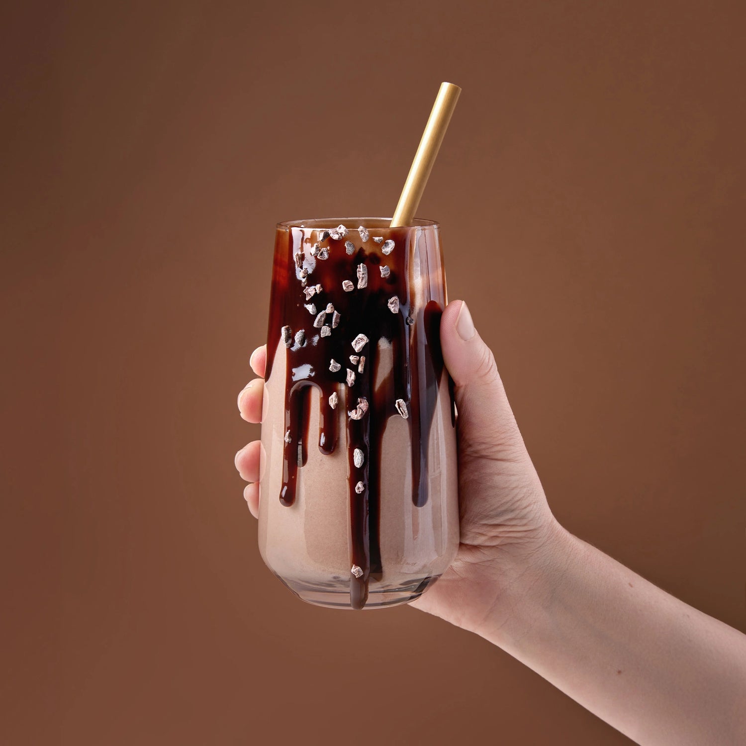 Chocolate meal replacement protein shake in a clear glass with chocolate drizzle and cacao nibs, gold straw, hand holding against a warm brown background