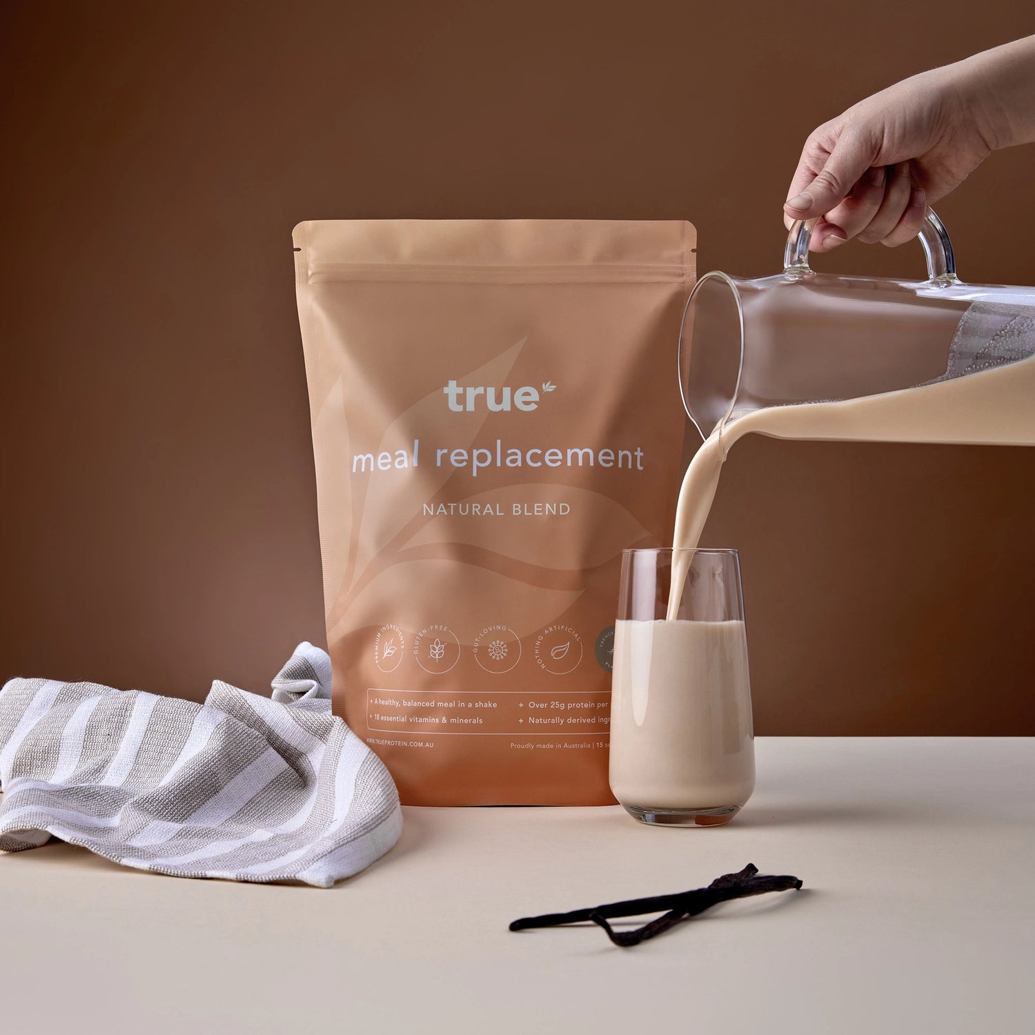 True Protein meal replacement natural blend pouch and vanilla-flavored shake being poured from a glass jug into a tumbler, with vanilla beans and striped kitchen towel on a neutral brown background.