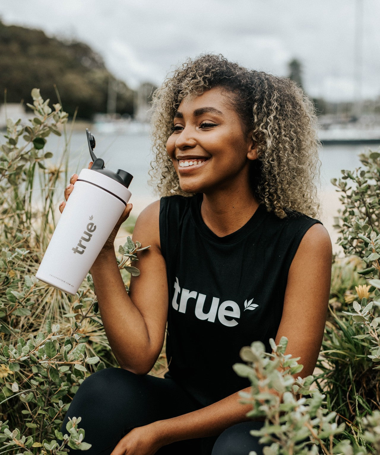 Smiling woman holding white &quot;True&quot; protein shaker bottle at a coastal marina in Clontarf, Australia — outdoor fitness lifestyle image