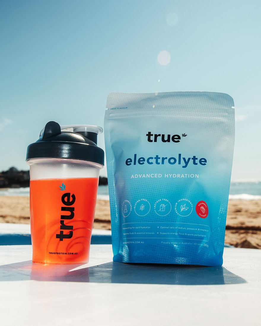 True Electrolyte 240g pouch in Strawberry beside a shaker of mixed drink on a surfboard at the beach