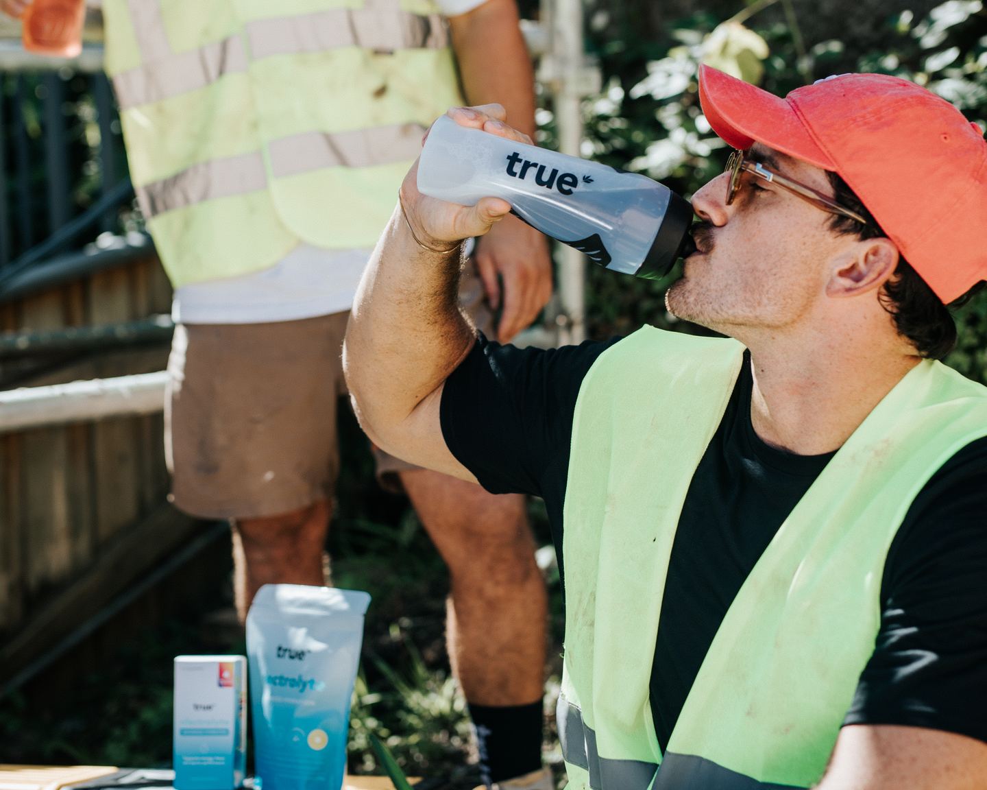 A tradie in a hi-vis vest drinks from a True drink bottle on site, with a True Electrolyte pouch and Travel Pack on a toolbox beside him