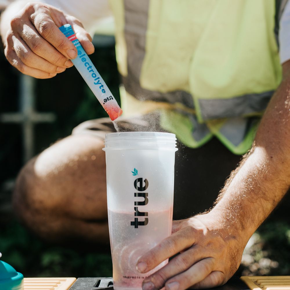 A tradie in hi-vis tears open a True Electrolyte sachet into a True shaker on a toolbox